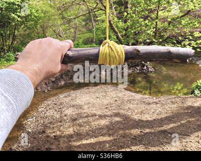 Close up details of a man holding onto a rope swing hanging from the branches of a mature tree in a forest. A small stick is tied with knot as a handle to swing over river below Stock Photo