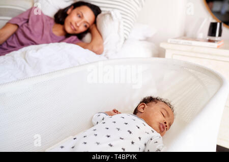 Close up of young adult mother lying on her bed looking down at her three month old baby sleeping in his cot Stock Photo