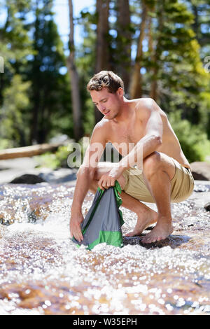 Camping hiking hiker man washing clothes on trek in nature river. Hiking young male adult doing clothing wash chores in natural stream of fresh water during an adventure trip outdoors. Stock Photo