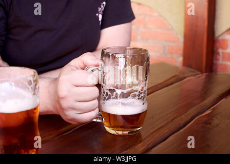 Man in t-shirt sitting at wooden table and holding in hand glass of beer with foam. He is thirsty and drinks alcohol. Stock Photo