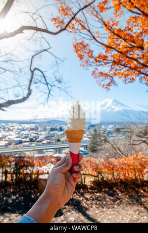 hand holding soft cream ice cream cone with Mount Fuji and colourful autumn maple tree background at Arakurayama Sengen Park - Fujiyoshida Stock Photo