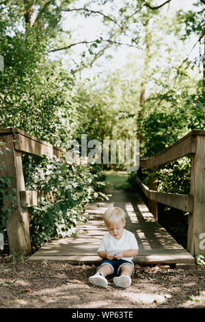 Cute Little Blond Haired Toddler Boy Kid Child Sitting and Laughing in Front of Wooden Bridge Over a Creek at the Park in the Forest During Summer Stock Photo