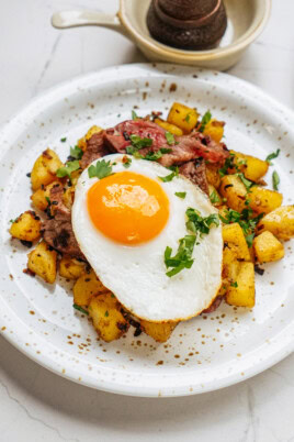 A plate of fried potatoes topped with chopped herbs, a slice of cooked meat, and a sunny-side-up egg. A small bowl sits in the background.