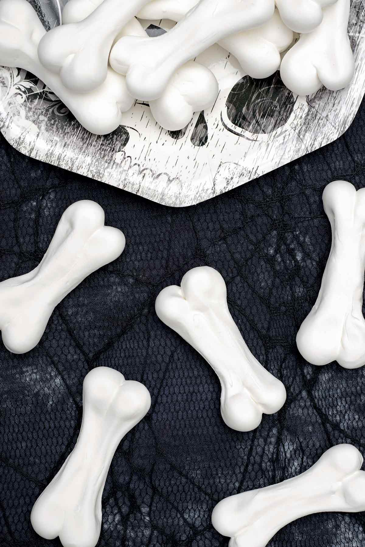 Halloween meringue bones piled on a paper plate and scattered on a black cobweb tablecloth beside the plate.