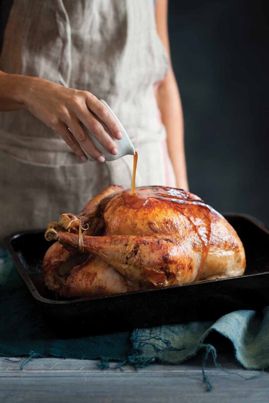 A woman pouring maple syrup over a maple glazed turkey in a black roasting pan.