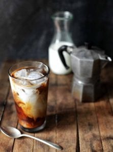 A glass of cold brew coffee with a French press and jar of cream in the background.