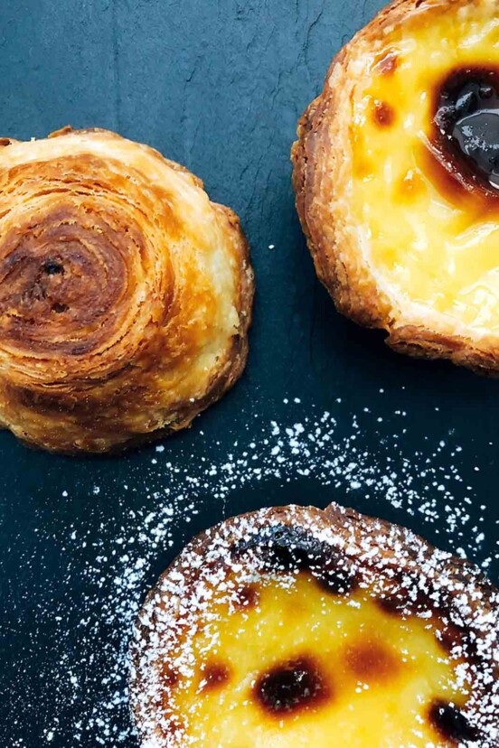 Three pasteis de nata on a slate background sprinkled with powder sugar.