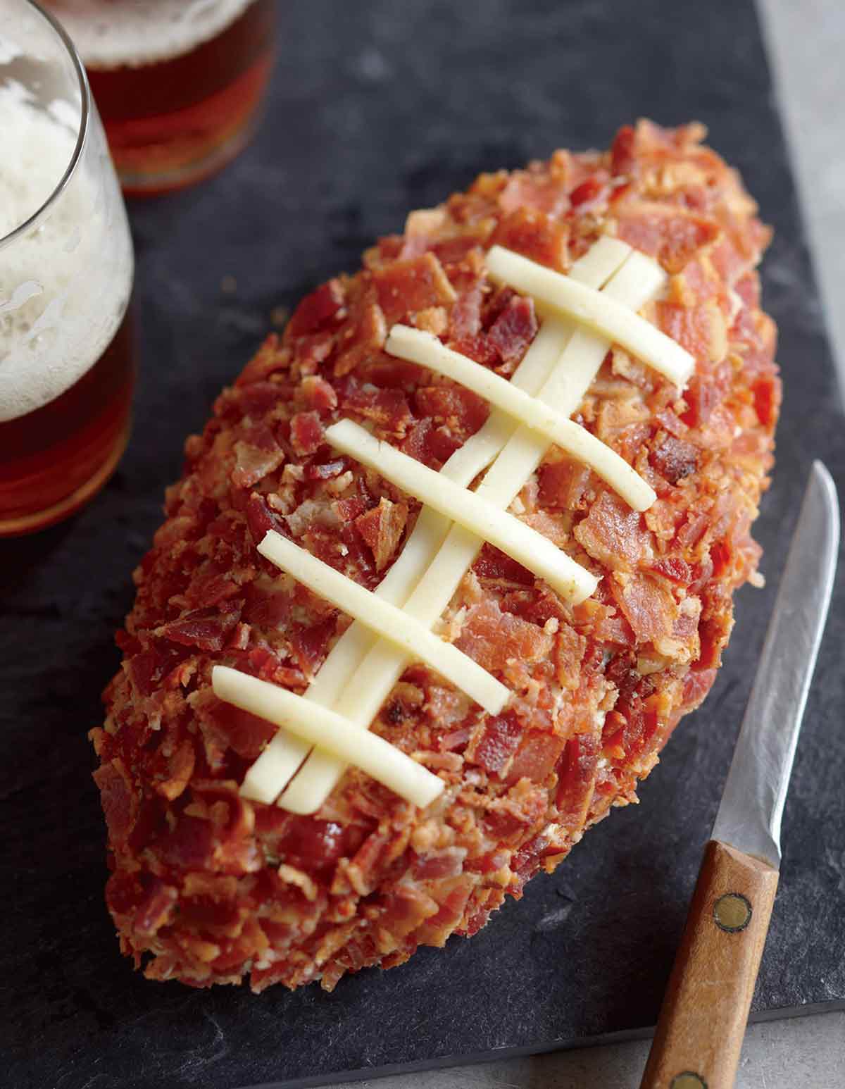 A football cheese ball on a serving board with two glasses of beer and a knife beside it.