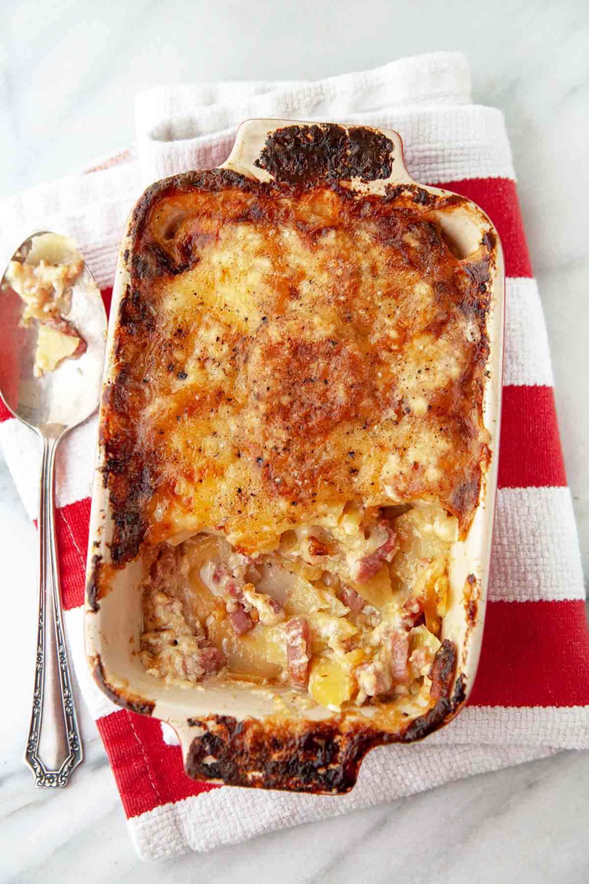 A ham, cheese, and potato casserole in a large rectangular dish, on a red-and-white striped towel.