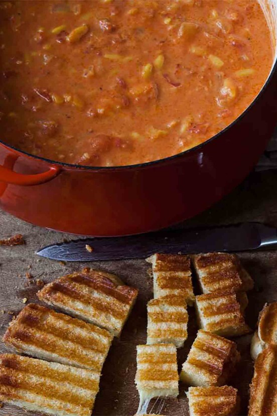 A large pot of Ina Garten's easy tomato soup with a grilled cheese sandwich being cut into croutons beside it.