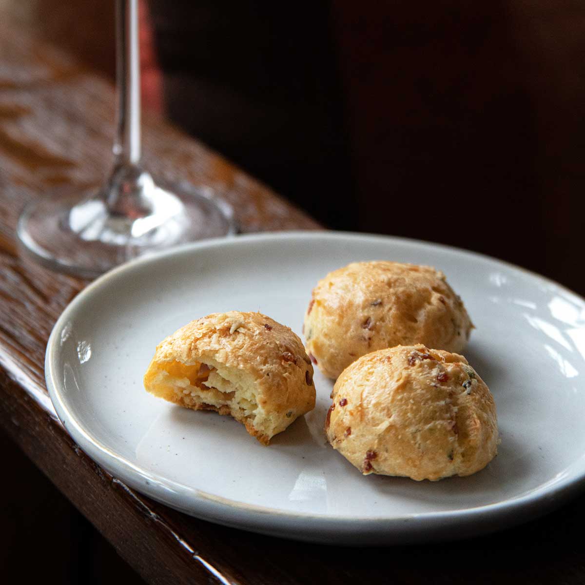 A white plate with three prosciutto-cheese gougeres with a glass of Prosecco in the background.