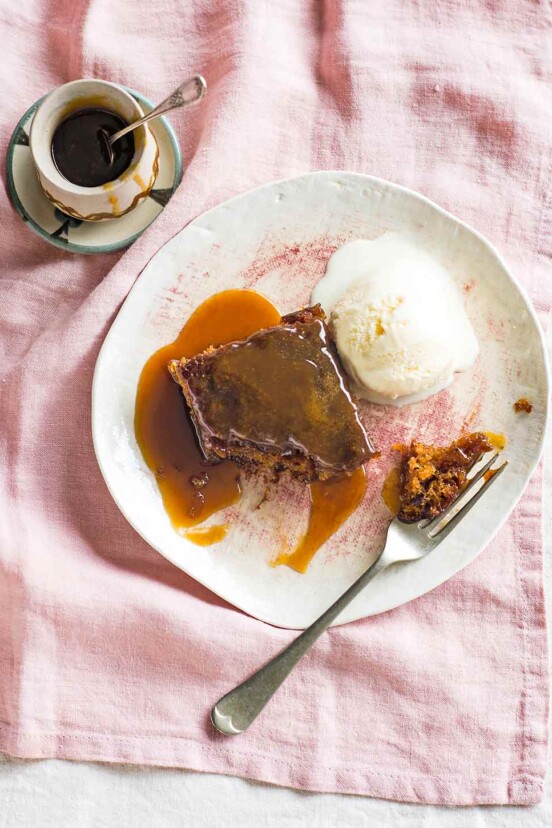 Sticky toffee pudding with vanilla ice cream on a white plate with a fork, beside a small bowl of extra toffee sauce.