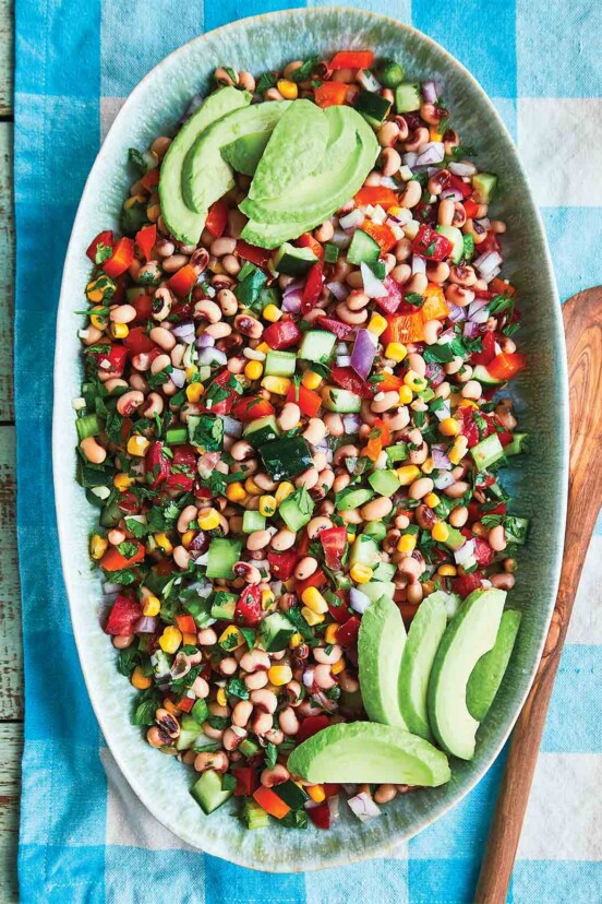 Black-eyed pea salad in a large oval bowl, on a blue and white checked tablecloth, beside a wooden spoon.