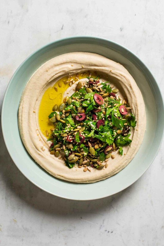 A pale blue bowl filled with white bean hummus, topped with parsley, olives, seeds, and oil