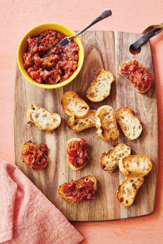 A wooden cutting board with a bowl of caramelized onion and tomato jam and several toasted baguette slices on it.