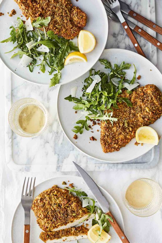 Three plates topped with pistachio-crusted chicken cutlets, arugula salad, and lemon wedges, with forks and knives on the side.