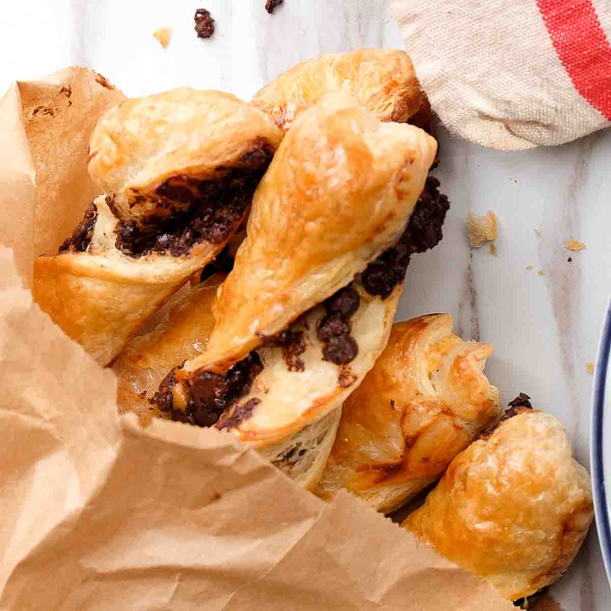 Torsades au chocolat in a brown paper bag on a marble counter, beside a white plate with pastry and a linen tea towel.