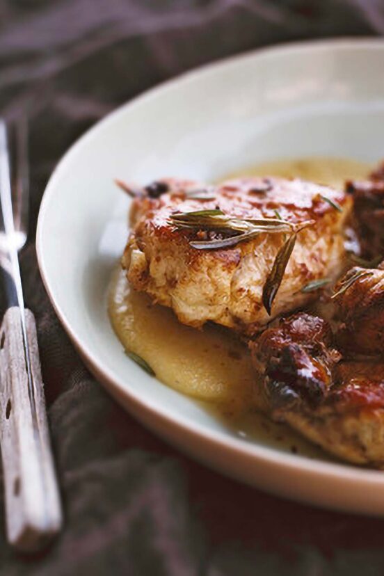 Chicken pieces in a sauce of vinegar, garlic, rosemary in a white plate, knife and fork on the side.