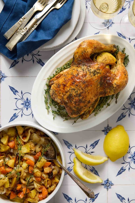 A whole roast chicken on a platter with a bowl of roasted root vegetables nearby.