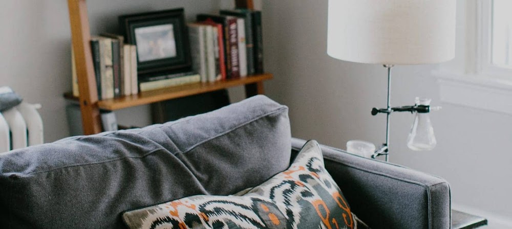 Close up photograph of a grey sofa in a living room with a lamp and wooden bookshelves.