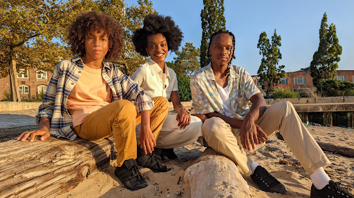 A young Black boy, a young Black woman, and a young Black man sit together in a park with the sun shining on them