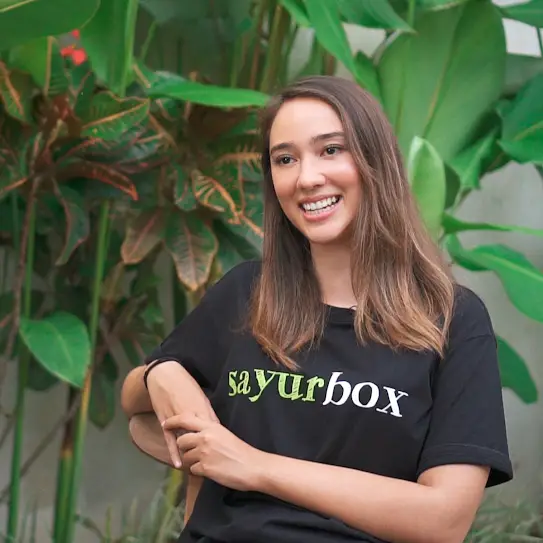 Seorang wanita mengenakan kaos berlogo Sayurbox, tersenyum ke arah kamera