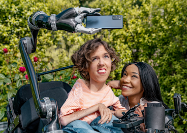 Une personne de type caucasien avec des cheveux bruns ondulés est assise dans un fauteuil roulant motorisé. Le fauteuil est équipé d'un bras robotisé qui tient un téléphone Android. Une femme noire est accroupie à côté et sourit en regardant le téléphone Android.