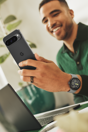 A young adult sitting at a desk smiles while looking at their Pixel 9 device.