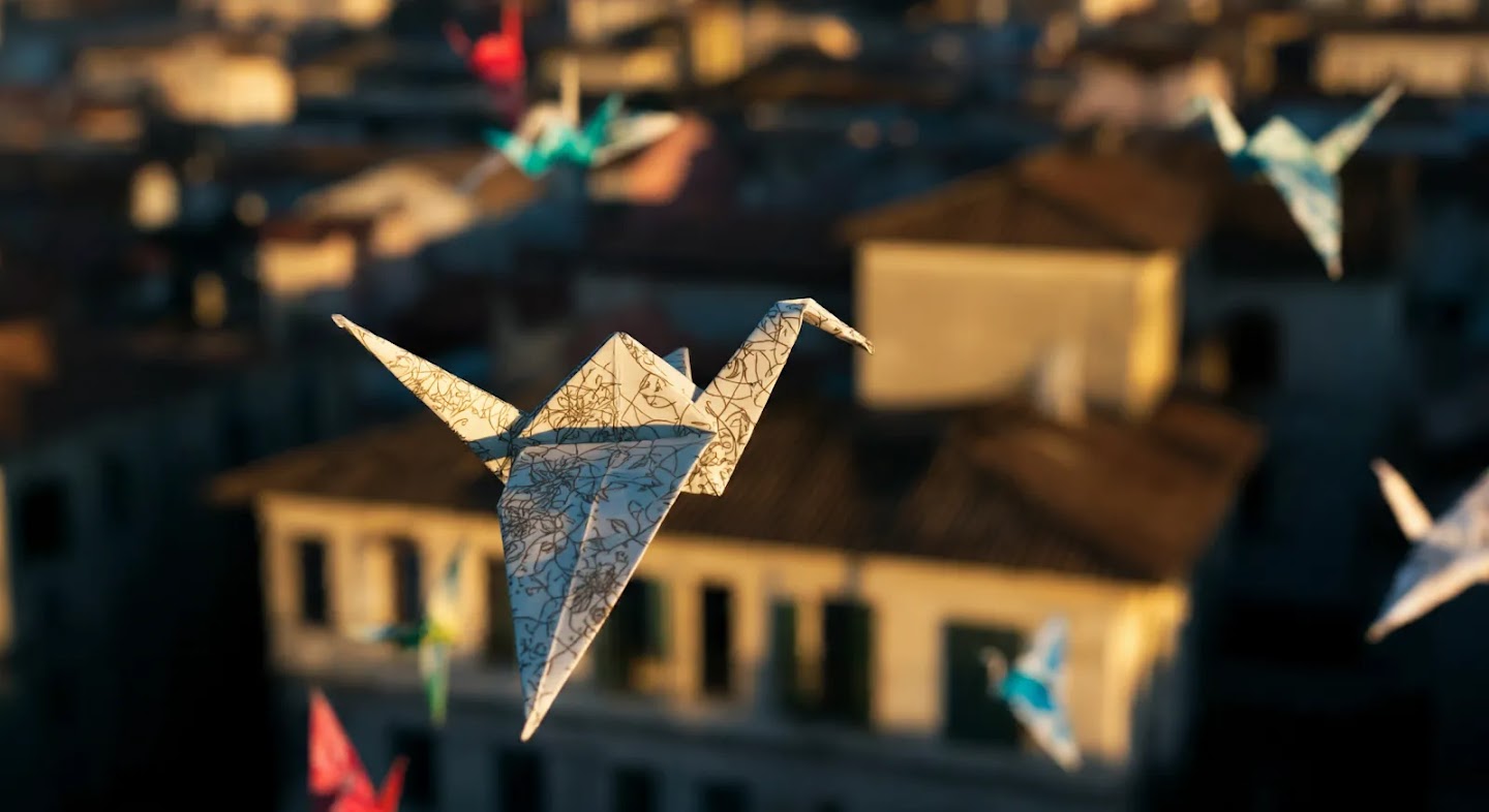 Numerous origami paper cranes, in various colors and patterns, float over a blurred cityscape with rooftops bathed in warm sunlight.