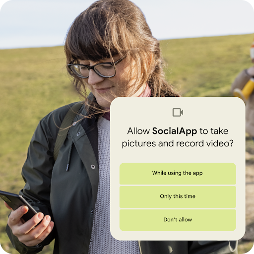 A person standing on a grassy hillslope looking at their Android phone. A graphic overlay seeking permission to allow an app to take pictures and record videos is placed on top. Permission options include allowing access while using the app, only this time or don’t allow.