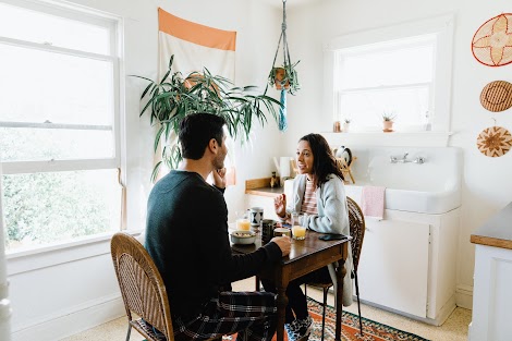 A man and woman in their pajamas talk at a small kitchen table and are undistracted by a phone.