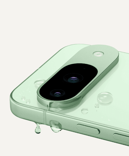 A close up of the top corner of Pixel 9 in Wintergreen color, one of its camera lenses in view. A single drop of water drips off the side of the phone.