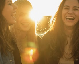 AI generated image of three women stand together laughing, with one woman slightly out of focus in the foreground. The sun is setting behind the women, creating a lens flare and a warm glow