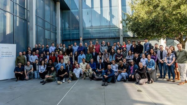 A large group of people stand in front of an office building in a tight group. A sign reading: Google.org Accelerator: Generative AI stands to the left of the group.