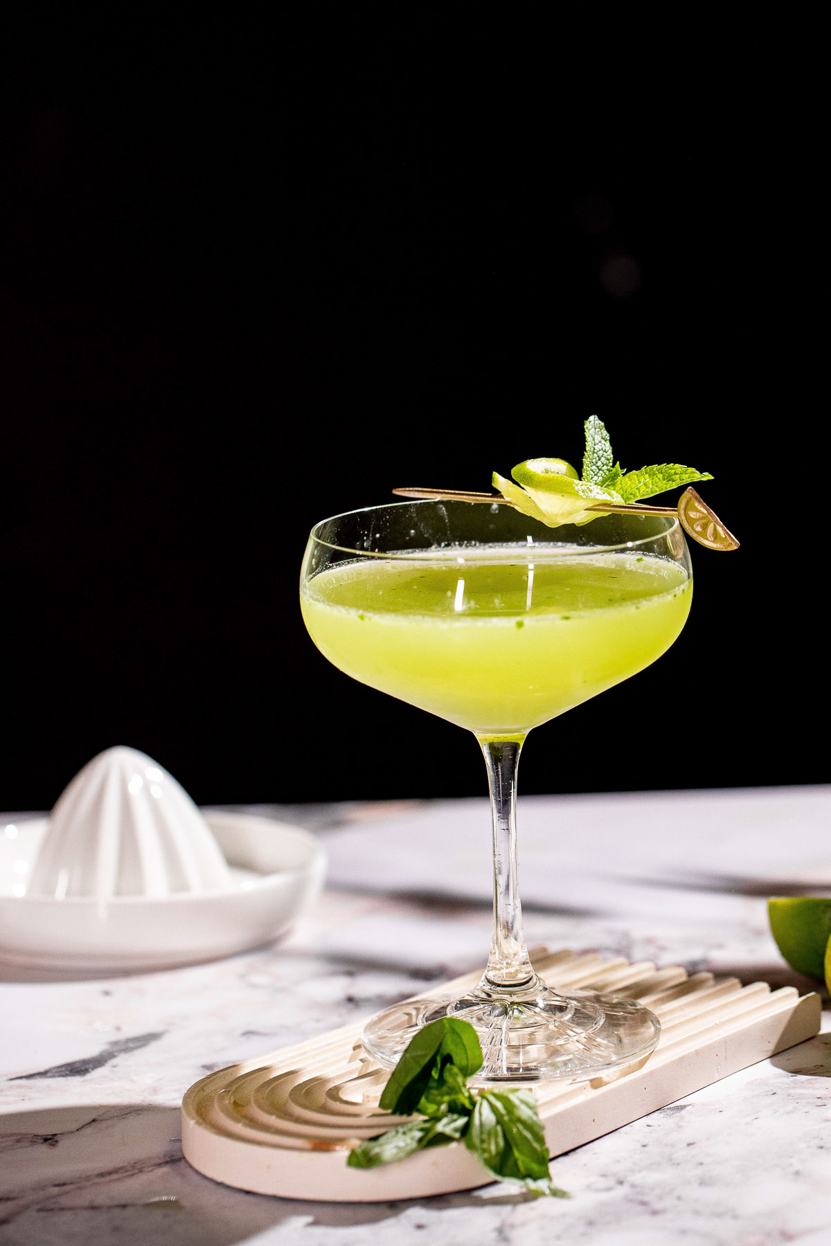 A basil martini garnished with lime twist and mint sits on a marble surface with a citrus juicer in the background.