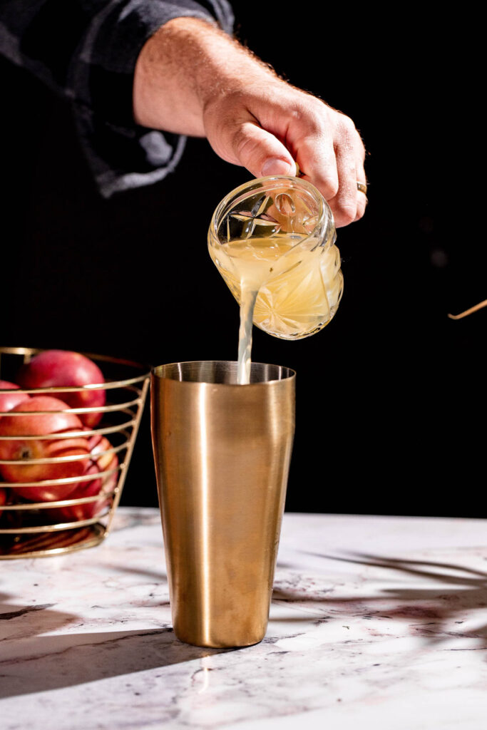 A person crafts an apple cider martini by pouring liquid into a metal shaker, with fresh apples in a wire basket nearby.