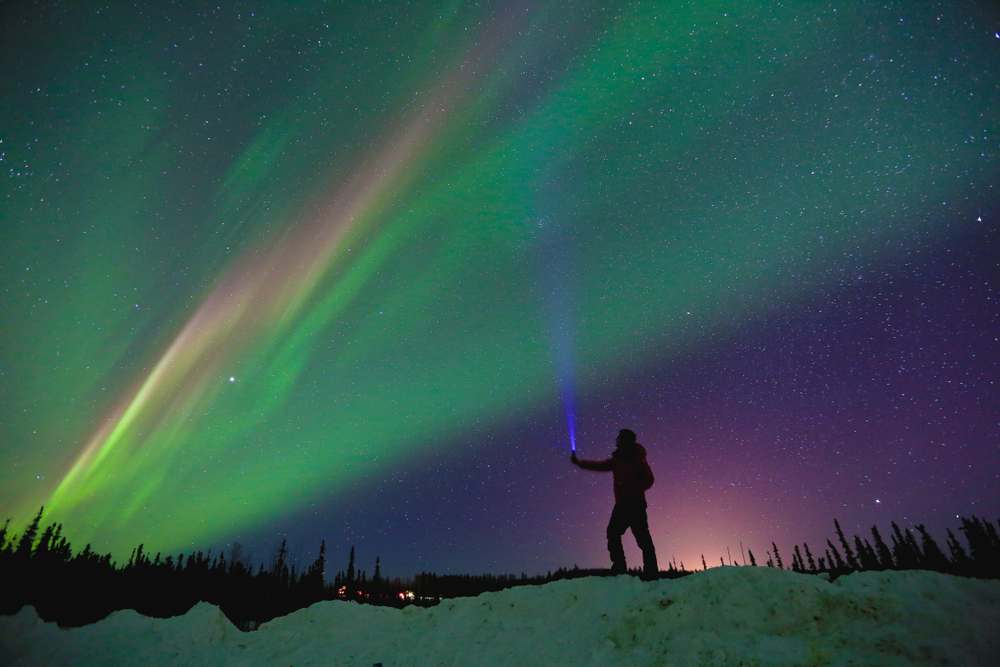 Person on snow covered hill shining a light towards the sky the northern lights are in the sky. 