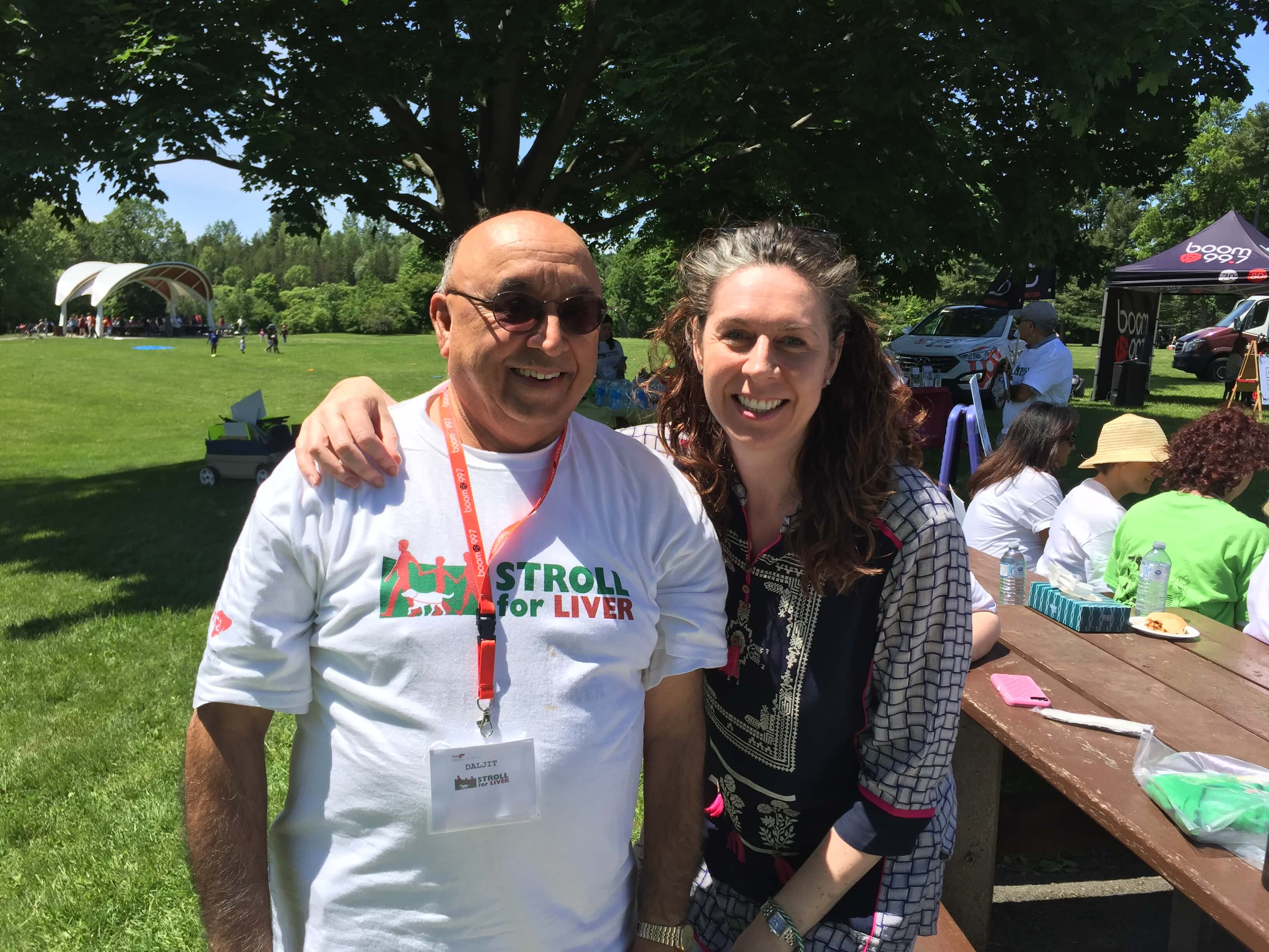 Daljit and Heather pose together smiling at the Ottawa Stroll for Liver. The background is a sunny day at a park, and they are positioned beside a picnic table.