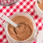 homemade cocoa mix in glass container on red checkered napkin with mug of cocoa