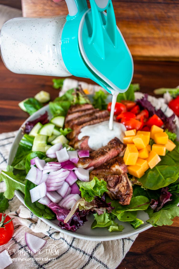 A vibrant steak salad with sliced steak, diced yellow cheese, red bell peppers, cucumbers, and onions is being drizzled with creamy dressing from a blue pitcher. The delightful mix sits on a wooden table beside a red striped napkin.