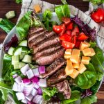 A steak salad featuring grilled steak slices, diced tomatoes, cubed cheddar cheese, red onion, and cucumber on a bed of mixed greens, garnished with fresh herbs. A striped cloth and scattered vegetables are artfully arranged in the background.