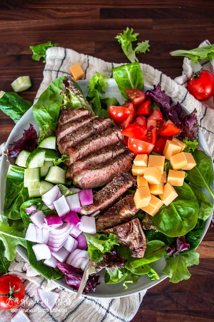 A steak salad featuring grilled steak slices, diced tomatoes, cubed cheddar cheese, red onion, and cucumber on a bed of mixed greens, garnished with fresh herbs. A striped cloth and scattered vegetables are artfully arranged in the background.