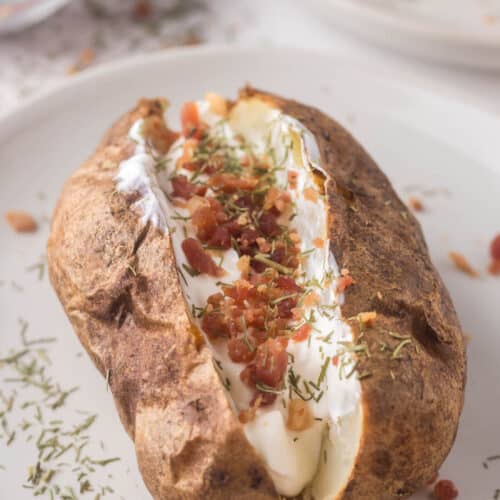 A baked potato, perfectly crisp from the best air fryer, is topped with sour cream, bacon bits, and herbs on a white plate. Additional bacon bits await in a small bowl in the background.