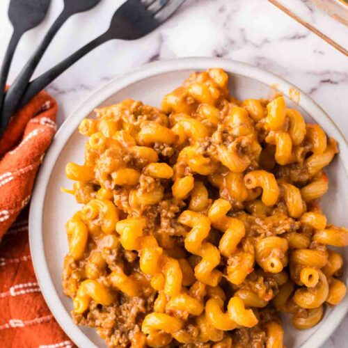 A plate of cheesy pasta with ground beef rests on a marble countertop, embodying an easy cheeseburger casserole recipe. The dish is flanked by an orange napkin and four black forks, while a glass baking dish brimming with more pasta sits temptingly in the background.