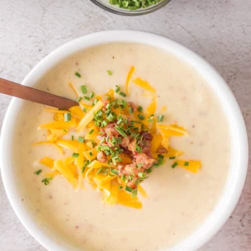 A bowl of creamy Dutch oven potato soup is topped with shredded cheddar cheese, chopped chives, and crumbled bacon. A spoon rests in the bowl, while a small dish of extra chives sits nearby.