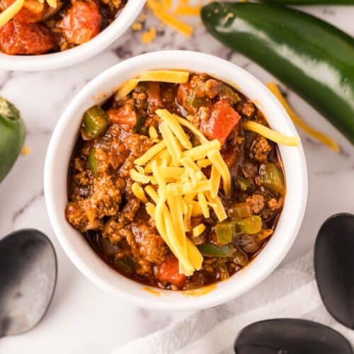 A bowl of the best Texas chili recipe topped with shredded cheese, surrounded by jalapeños and black spoons on a marble surface. The chili, featuring ground meat, tomatoes, and green peppers, sits enticingly next to another partially visible bowl.