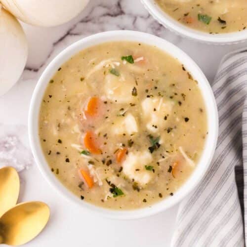 A bowl of creamy chicken and vegetable soup crowned with fluffy Bisquick dumplings is garnished with herbs, sitting on a marble countertop. Nearby, two white onions and a striped cloth napkin with two golden spoons complete the cozy setting.