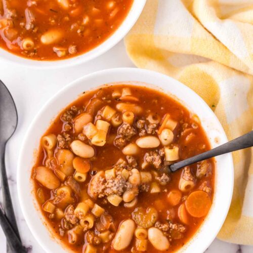 A bowl of hearty minestrone soup filled with white beans, ditalini pasta, ground meat, and carrots in a tomato broth. Inspired by an easy pasta fagioli soup recipe, another bowl is partially visible above, next to a folded yellow and white checkered napkin with two black spoons nearby.