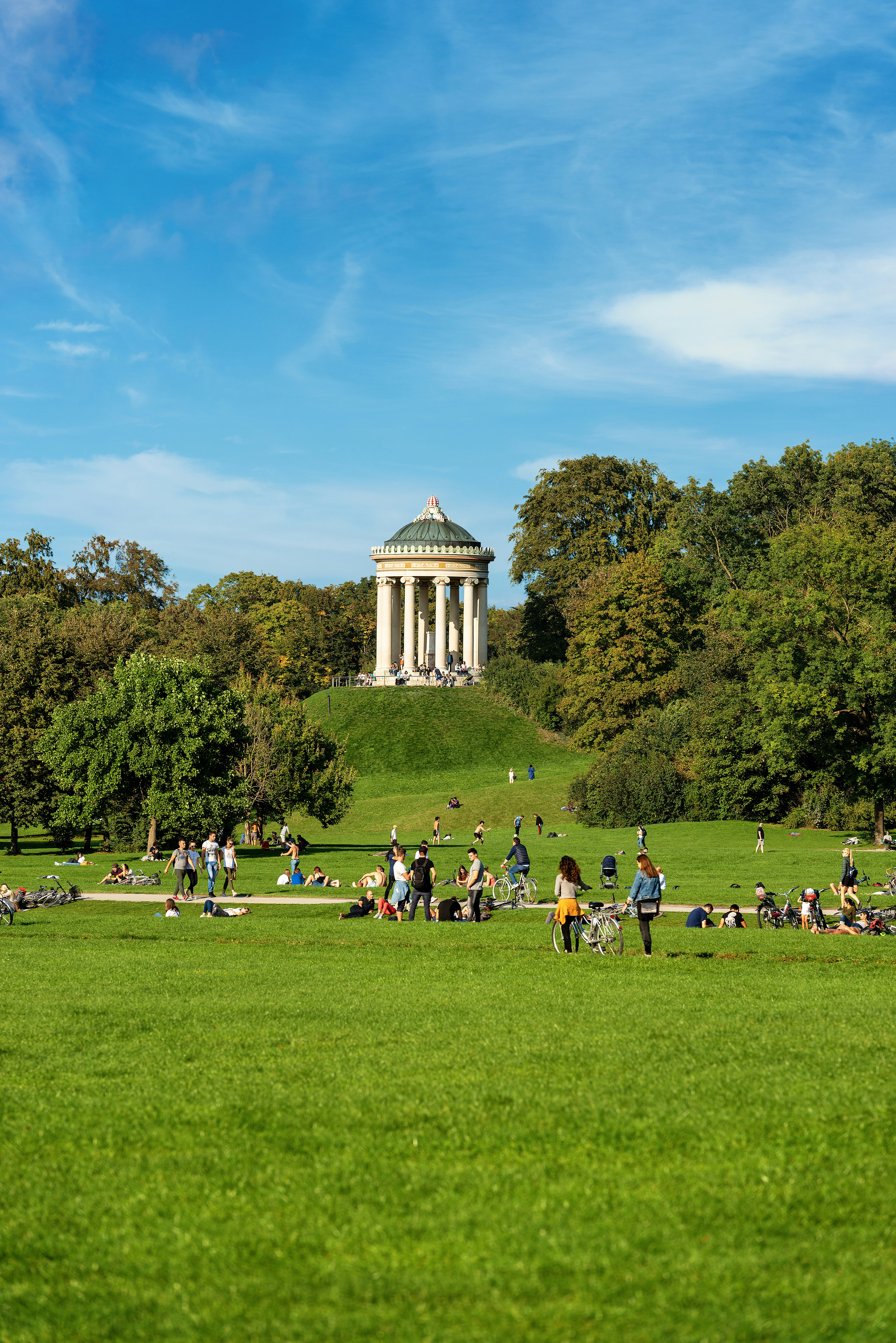 People sit on grass. A small round white temple-like building stands at the top of a hill