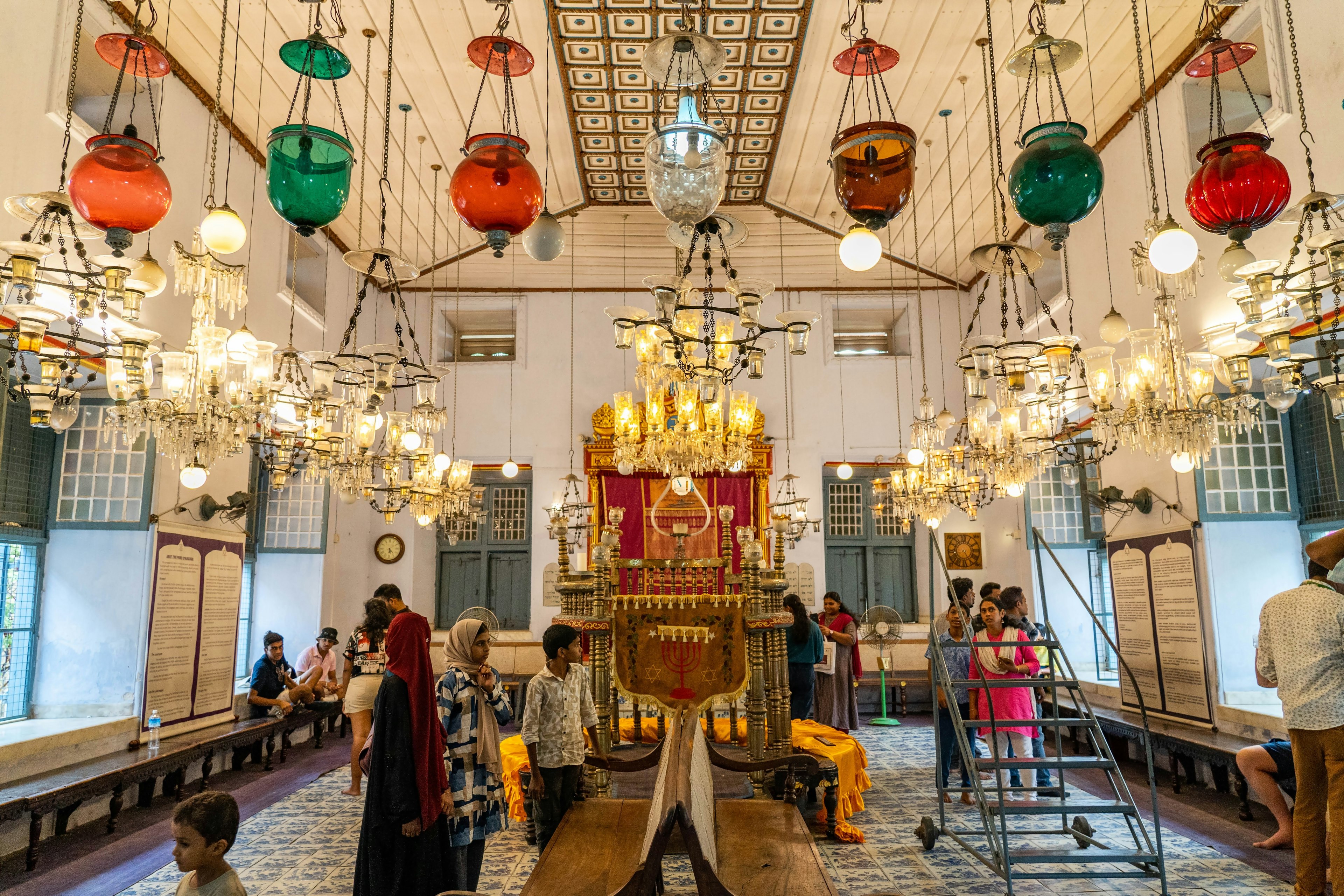 The interior of a synagogue featuring glass globe lanterns, chandeliers, and tiled floors. Kochi, India.